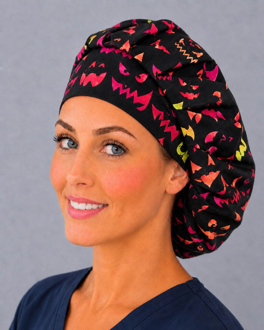 Woman wearing a neon pumpkin Halloween bouffant scrub cap — hot pink and orange jack-o'-lantern faces on black, adjustable women's surgical hat