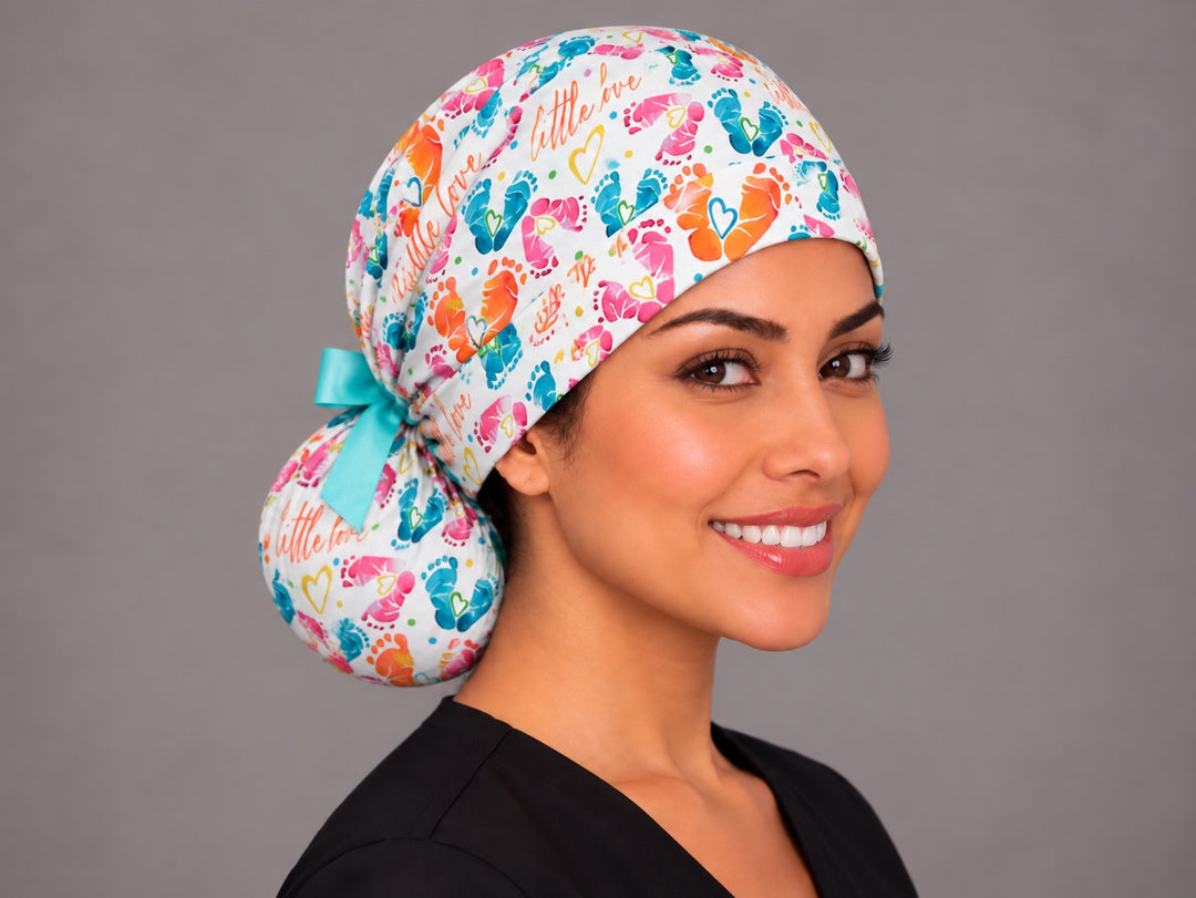 Right three-quarter angle of an OB nurse ponytail scrub hat, with large orange and pink watercolor footprints across the gathered crown
