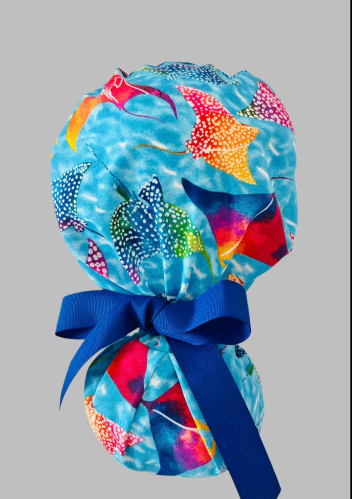 Ocean stingray ponytail scrub cap back view close-up showing gathered pouch full of colorful rainbow manta rays on blue water and navy ribbon bow