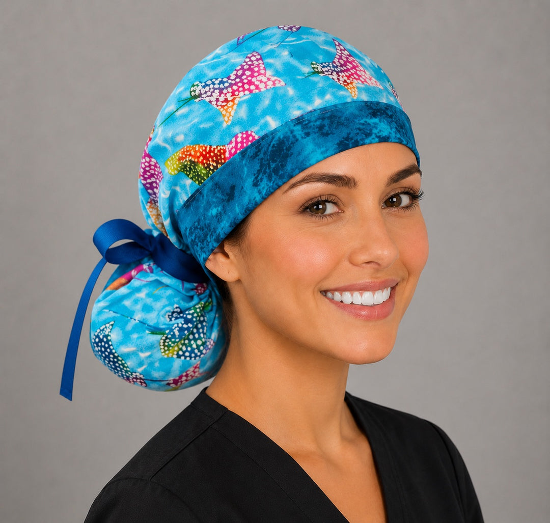 Ocean stingray ponytail scrub cap front-left angle on smiling model showing bright rainbow manta rays swimming on blue water print with navy band