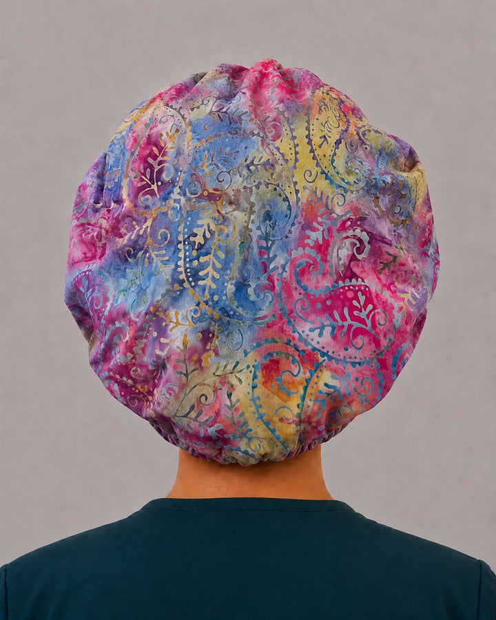 Woman wearing a pastel batik bouffant scrub cap, front view — hand-dyed fuchsia, powder blue and lavender paisley, adjustable women&