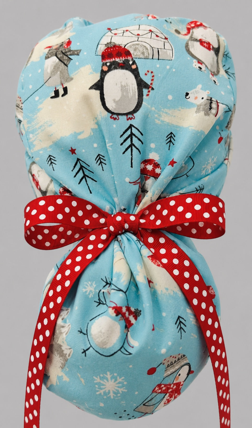 Woman wearing penguins and igloos ponytail scrub cap, front view smiling with penguins in red scarves, igloos and snowflakes on light blue with white snowflake band and red polka-dot ribbon tie