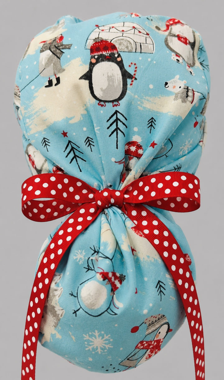 Woman wearing penguins and igloos ponytail scrub cap, front view smiling with penguins in red scarves, igloos and snowflakes on light blue with white snowflake band and red polka-dot ribbon tie