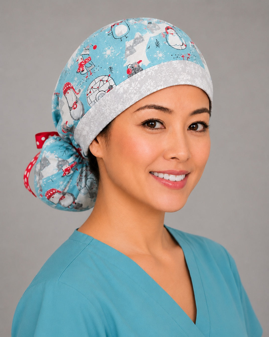 Penguins and igloos ponytail scrub cap left side profile on smiling model showing penguins, igloos and polar bears on light blue with white snowflake band and red polka-dot ribbon