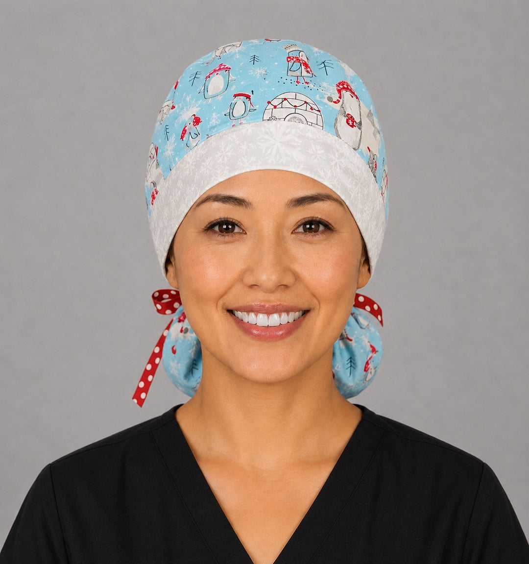 Penguins and igloos ponytail scrub cap left side profile showing gathered ponytail pouch of penguins, igloos and snowflakes on light blue with snowflake band and red polka-dot ribbon tie