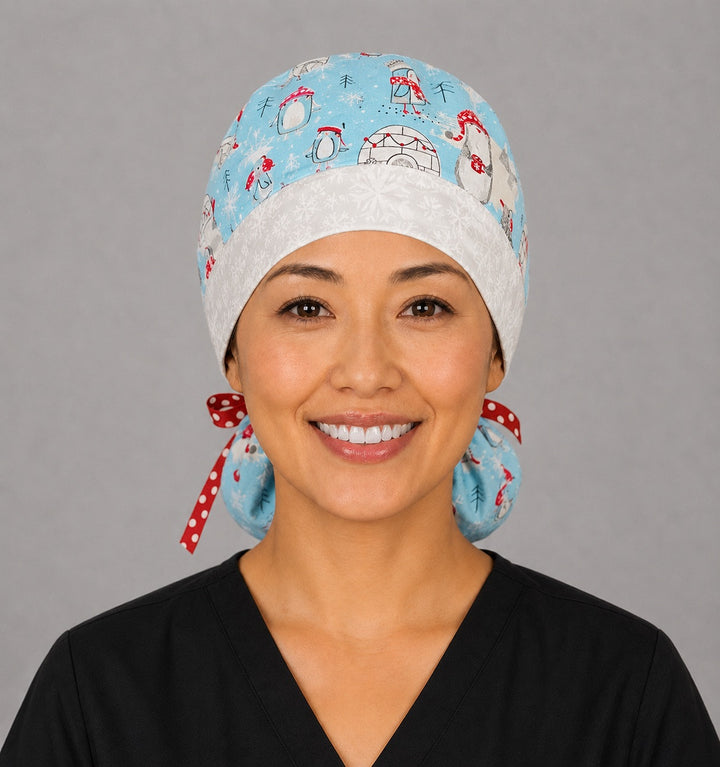 Penguins and igloos ponytail scrub cap left side profile showing gathered ponytail pouch of penguins, igloos and snowflakes on light blue with snowflake band and red polka-dot ribbon tie