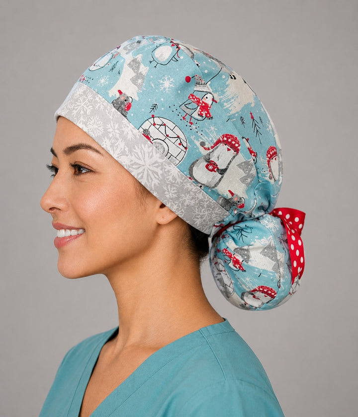 Penguins and igloos ponytail nurse cap right three-quarter angle on smiling model showing penguins in red scarves, igloos and polar bears on light blue with snowflake band and red polka-dot ribbon bow