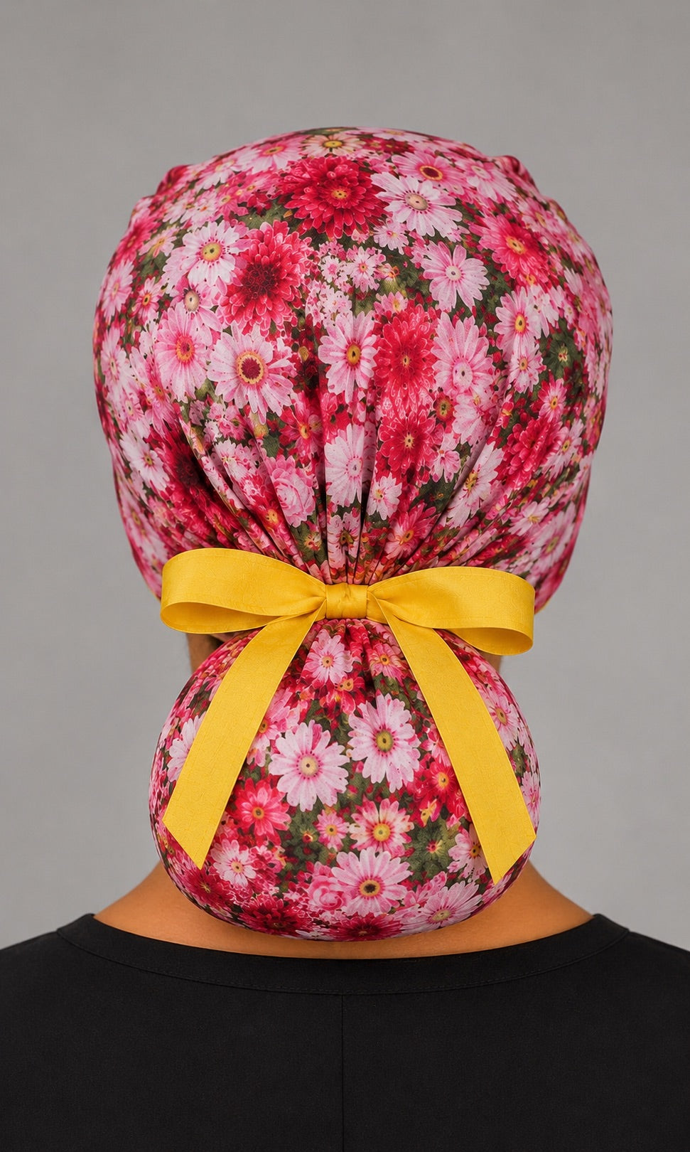 Woman wearing pink daisy floral ponytail scrub cap, front view smiling with pink daisies, roses and yellow band and ribbon tie