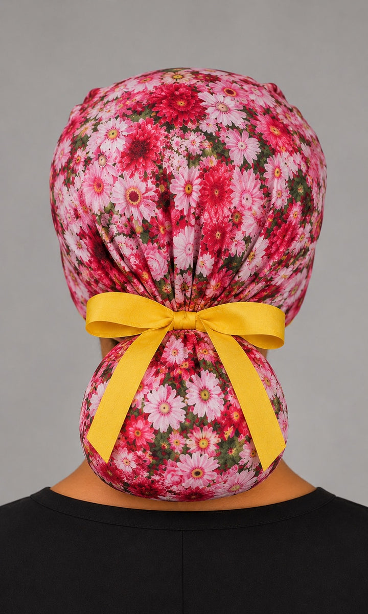 Woman wearing pink daisy floral ponytail scrub cap, front view smiling with pink daisies, roses and yellow band and ribbon tie