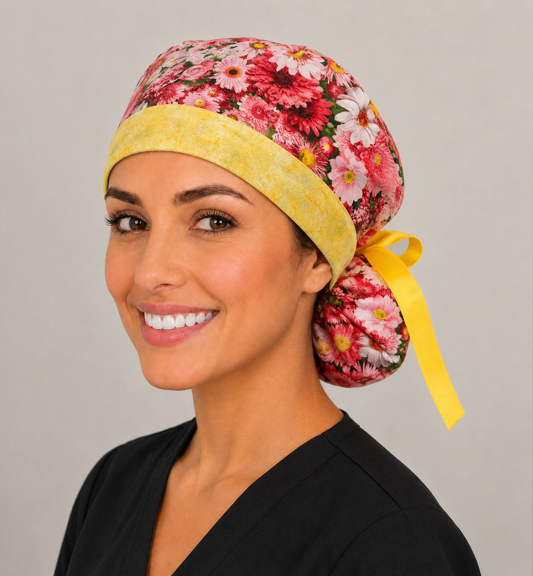 Pink daisy floral ponytail scrub cap right front angle on smiling model showing pink daisies, roses and yellow band with ribbon tie