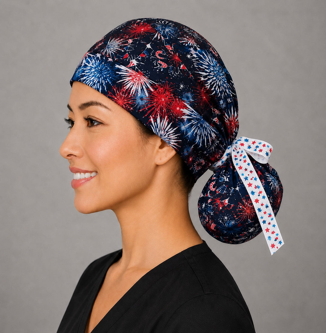 Profile view of model wearing navy patriotic fireworks ponytail scrub cap with USA print and white star ribbon tie