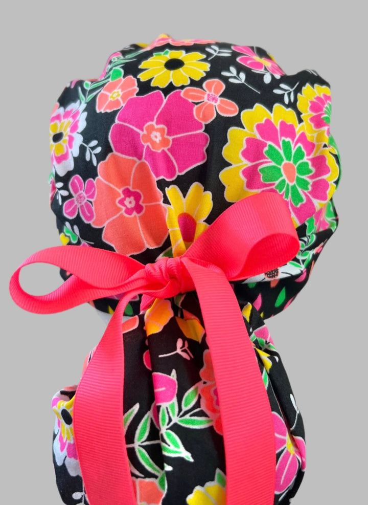 Close-up back view of neon floral ponytail scrub cap on a stand showing tied pink ribbon bow and gathered pouch