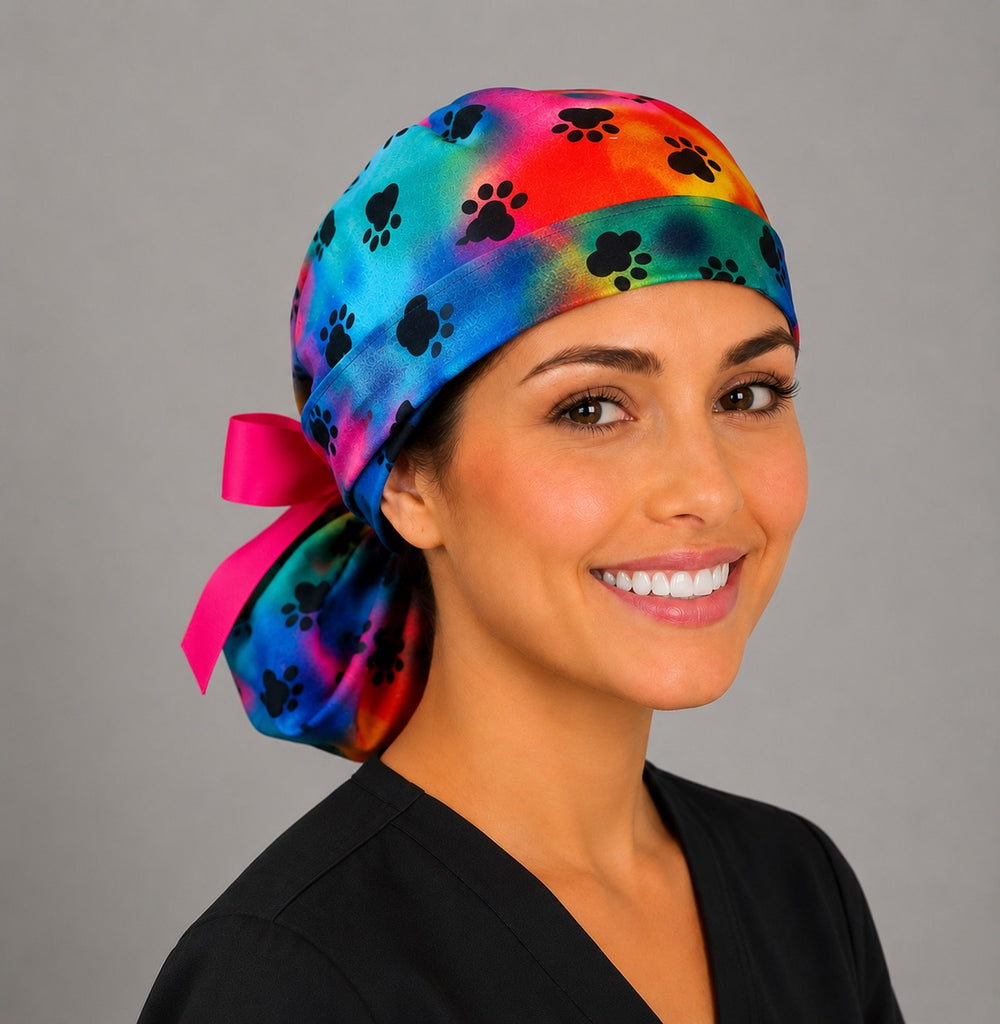 Woman wearing tie dye puppy paw rainbow ponytail scrub cap, front three-quarter view with black paw prints and hot pink ribbon bow