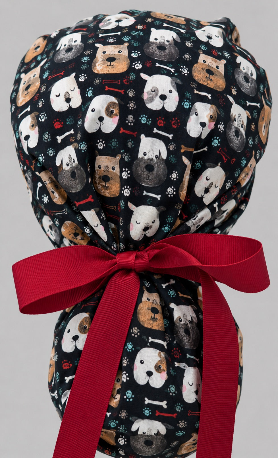 Woman wearing puppy dog lover ponytail scrub cap, front view smiling with cartoon dog faces, bones, paw prints on black and red ribbon tie