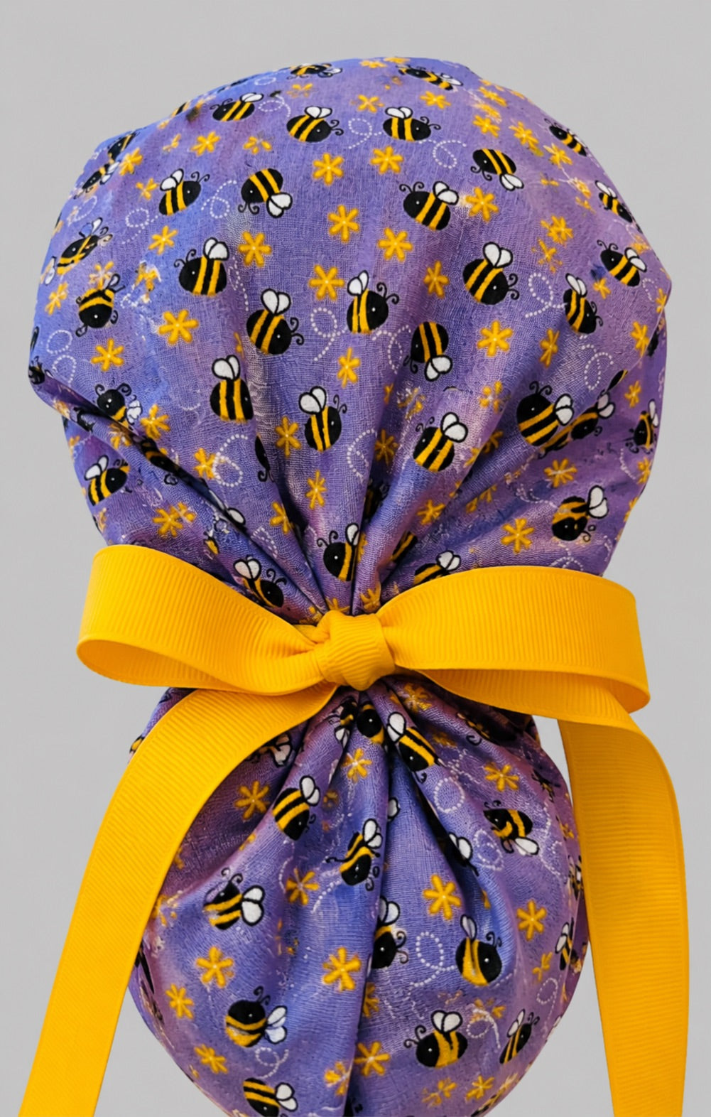 Purple bee and daisy ponytail scrub cap back view close-up showing gathered pouch of bumblebees on purple and yellow ribbon bow