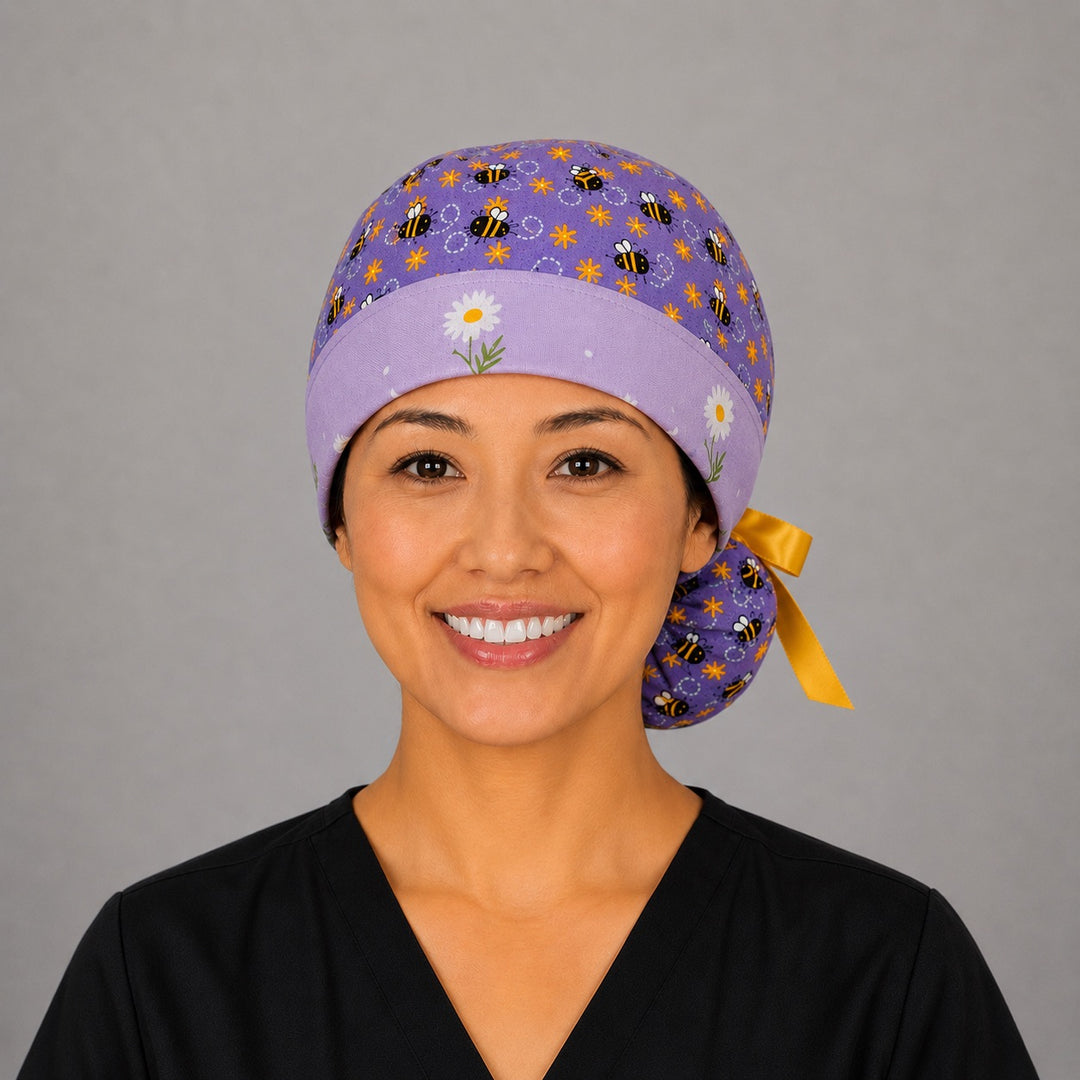 Woman wearing purple bee and daisy ponytail scrub cap, front view smiling with bumblebees, daisy band and yellow ribbon tie
