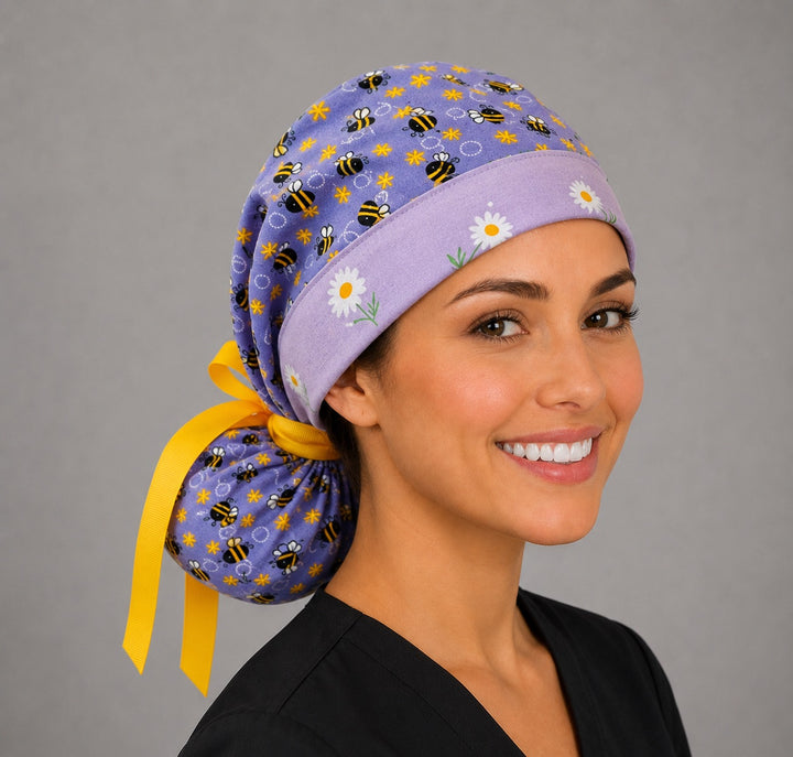 Purple bee and daisy ponytail scrub cap left front angle on smiling model showing bumblebees, daisies and yellow ribbon tie-back