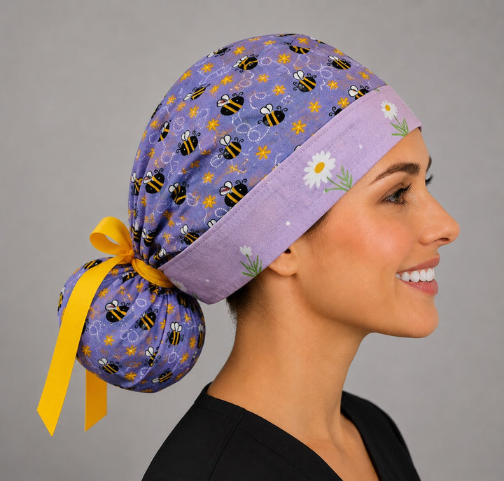 Purple bee and daisy ponytail scrub cap left three-quarter angle showing full ponytail pouch of bumblebees and yellow ribbon bow