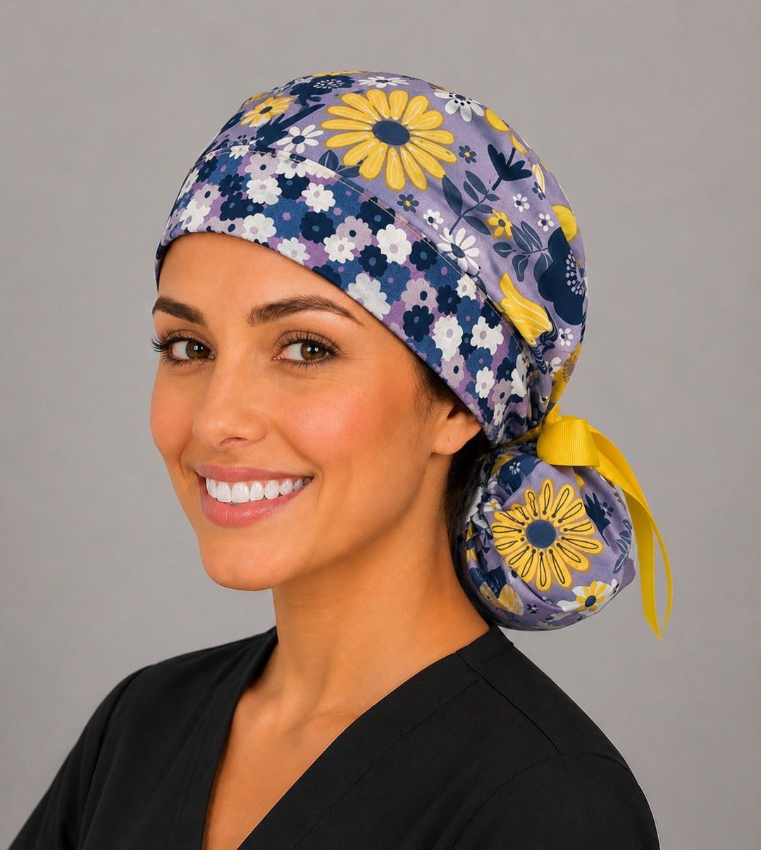 Purple and yellow floral ponytail scrub cap front-left angle on smiling model showing lavender daisies, yellow flowers and yellow ribbon tie