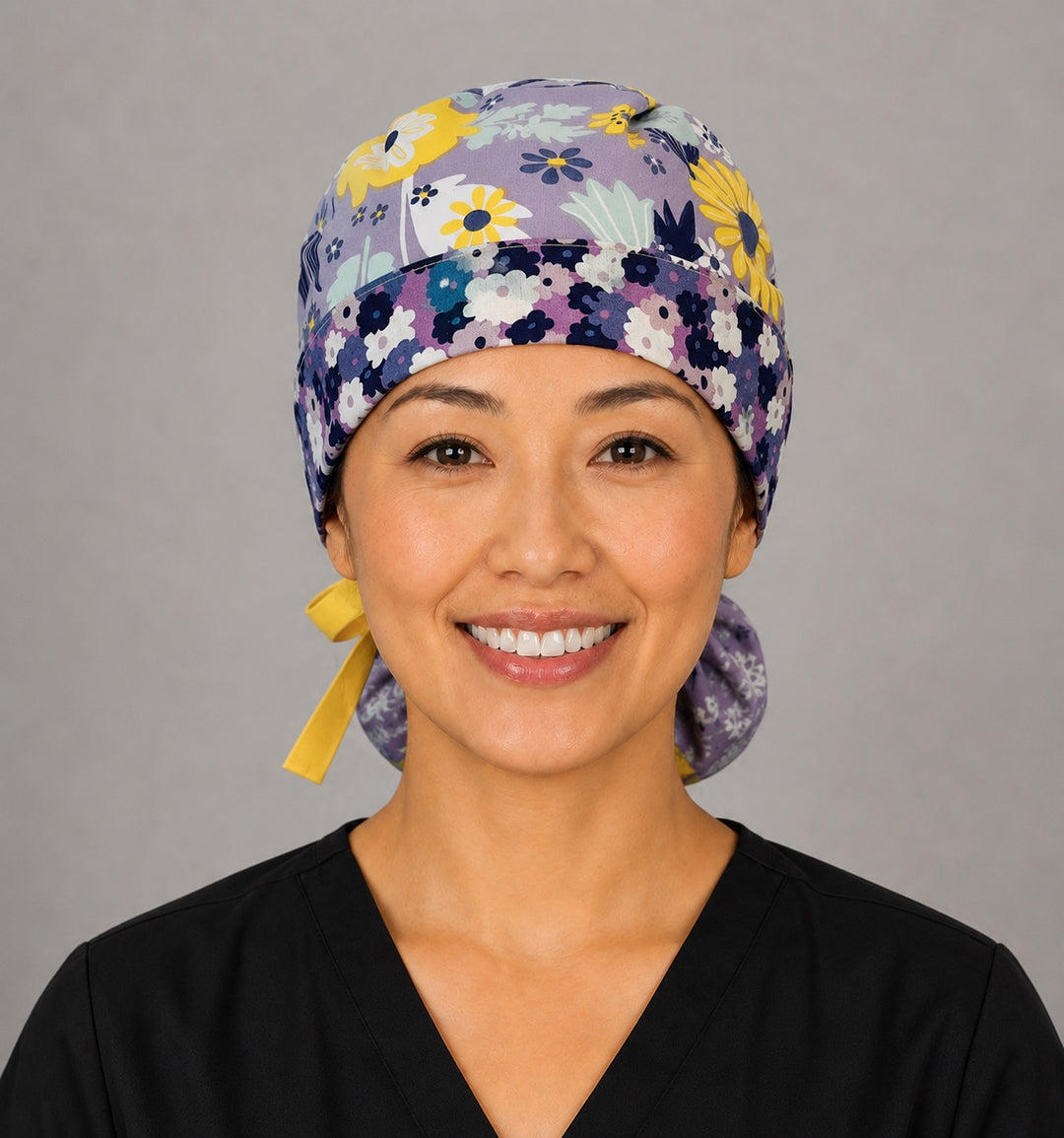Woman wearing purple and yellow floral ponytail scrub cap, front view smiling with lavender, navy and yellow summer flowers and yellow ribbon tie