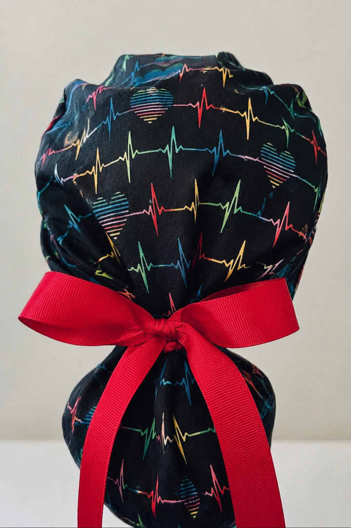 Rainbow EKG heartbeat ponytail scrub cap back view close-up showing gathered pouch full of colorful heartbeat lines and hearts on black with red ribbon bow