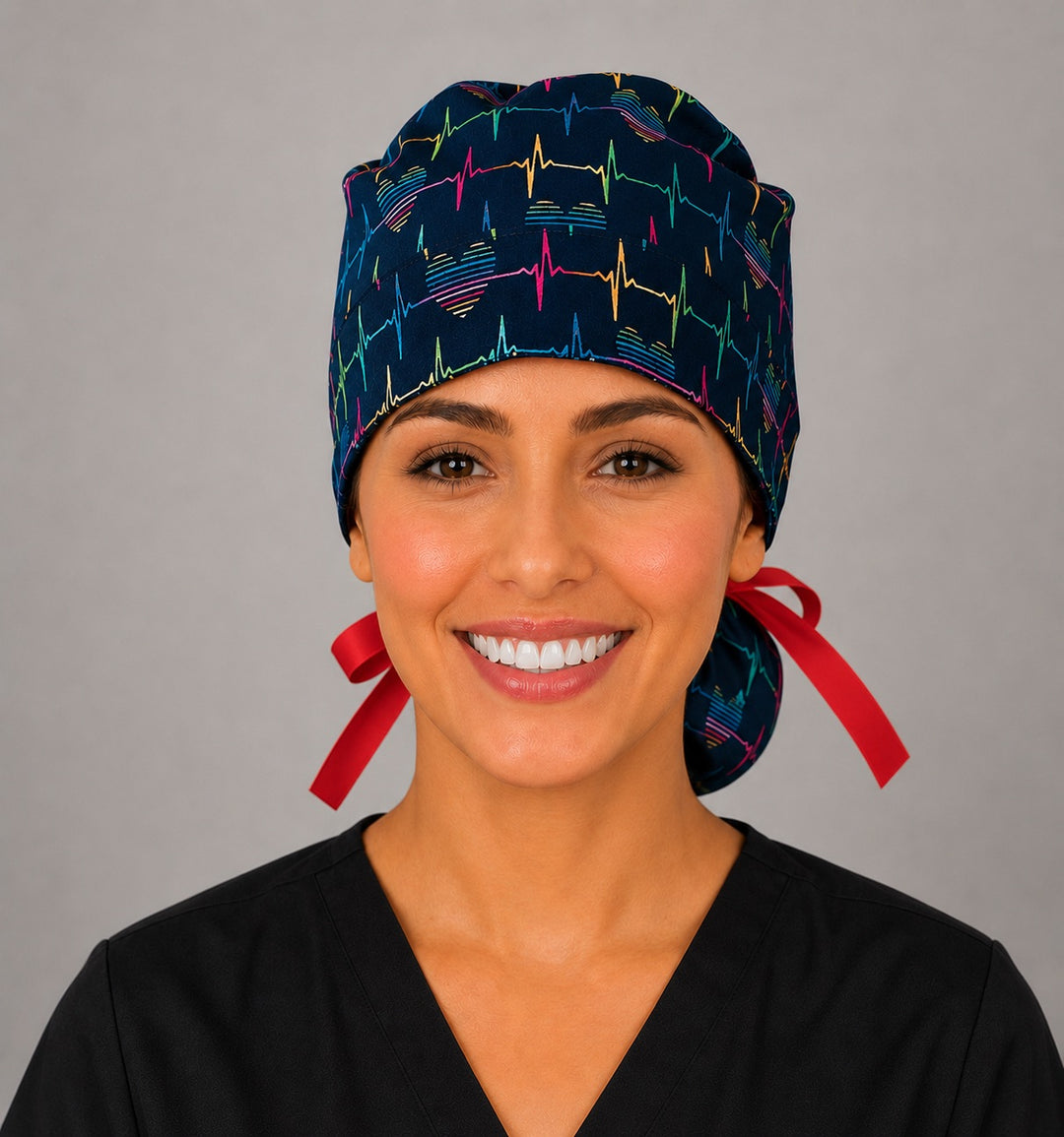 Woman wearing rainbow EKG heartbeat ponytail scrub cap, front view smiling with colorful heartbeat lines and hearts on navy with red ribbon tie