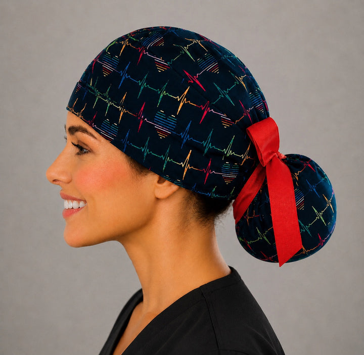 Rainbow EKG heartbeat ponytail scrub cap left side view on smiling model showing colorful heartbeat lines, hearts on navy and red ribbon bow at gathered pouch