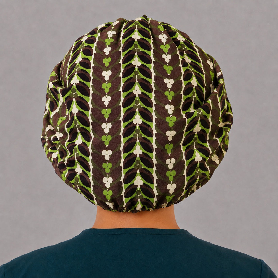 Woman wearing a retro leaf bouffant scrub cap, front view — chocolate brown and green vintage leaf print with cream clovers, adjustable women&
