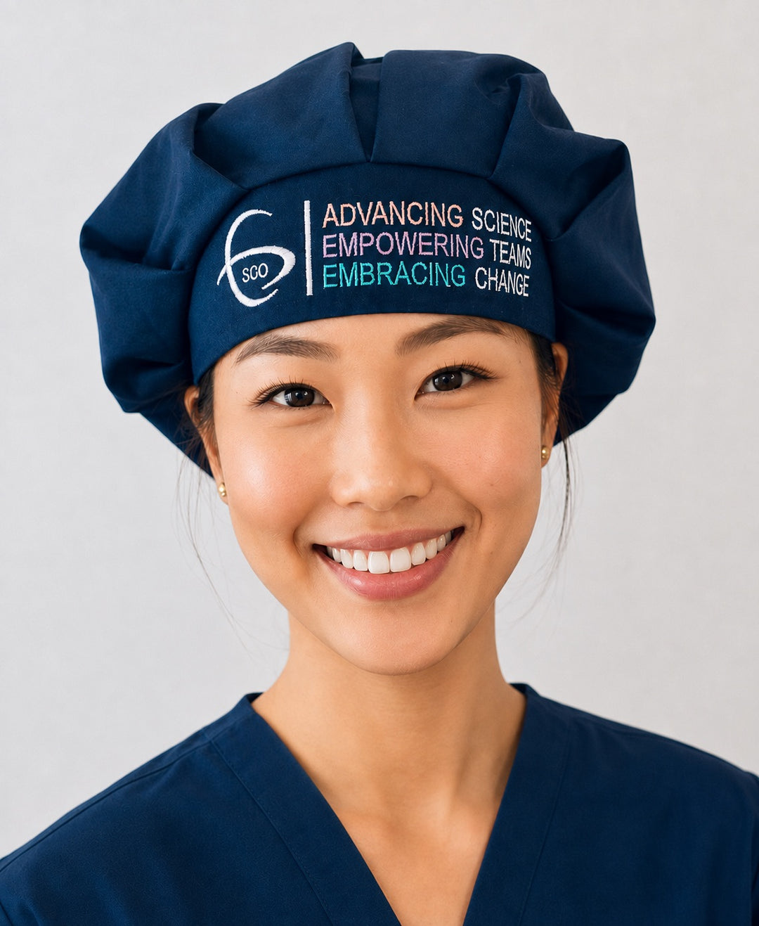 Smiling OR nurse in a navy SGO official embroidered bouffant surgical scrub cap with the full-color Society of Gynecologic Oncology logo across the front band