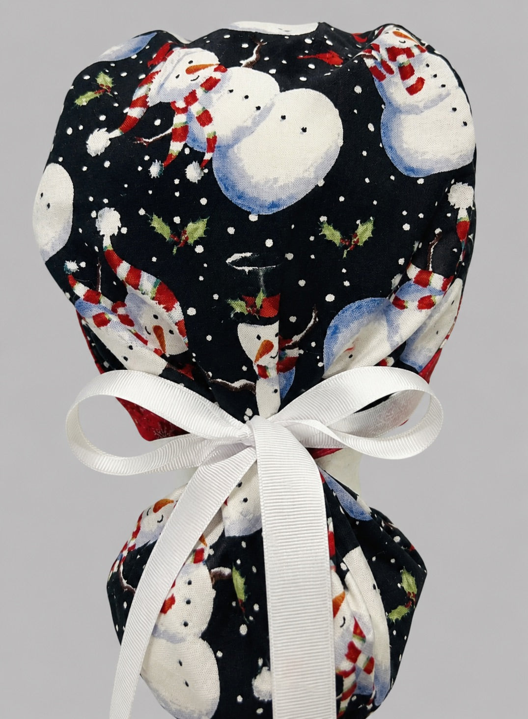 Woman wearing snowmen ponytail scrub cap, front view smiling with snowmen in striped hats and scarves on black with red snowflake band and white ribbon tie