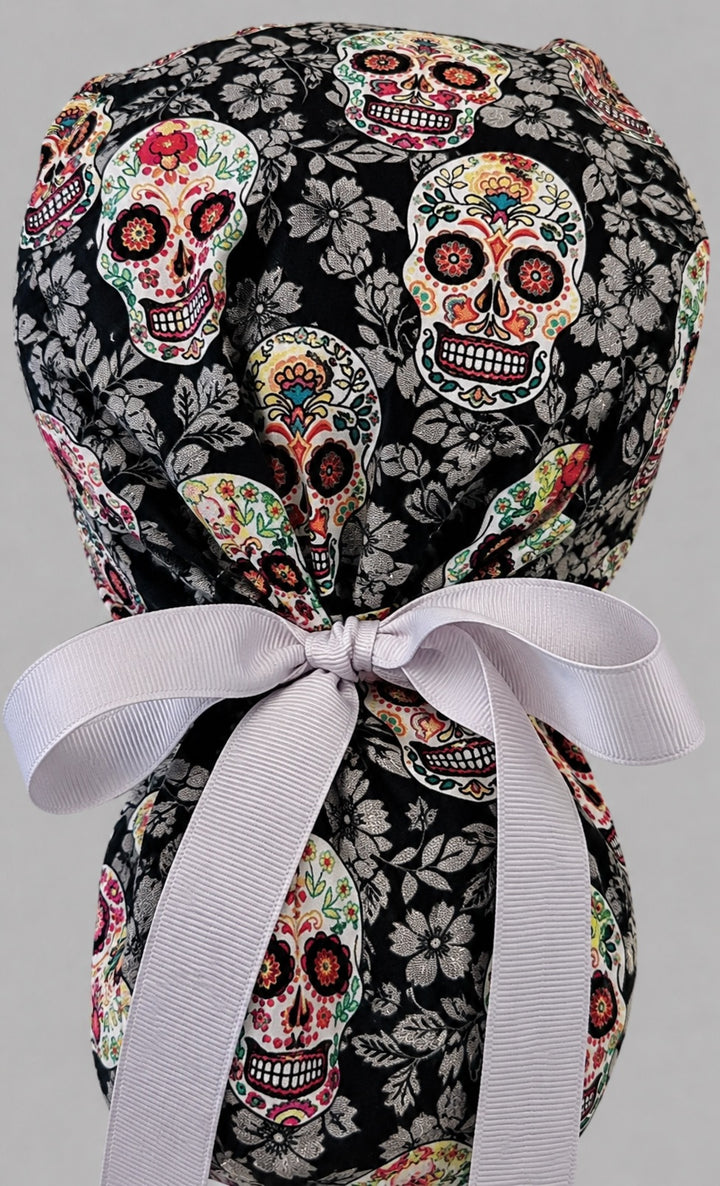 Sugar skull Halloween ponytail scrub cap back view close-up showing gathered pouch full of colorful Day of the Dead skulls on black floral print with gray grosgrain ribbon bow