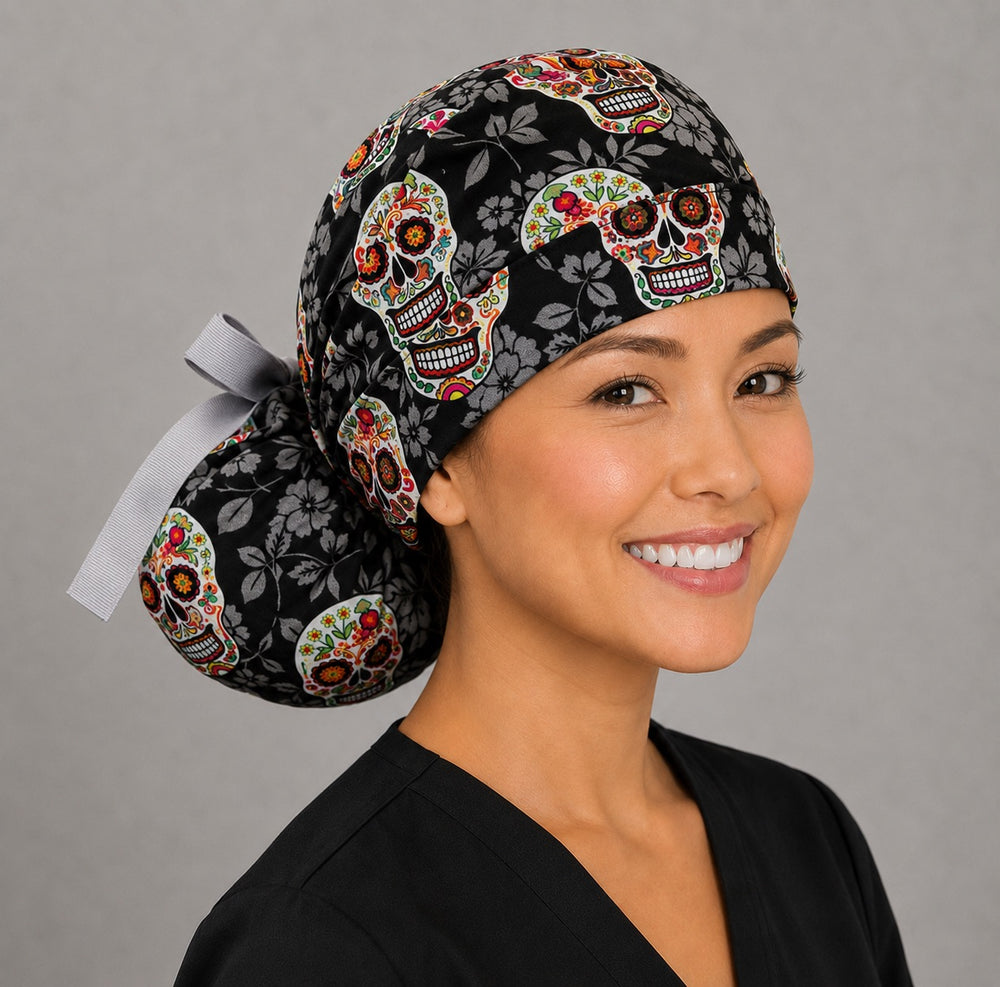 Woman wearing sugar skull Halloween ponytail scrub cap, front view smiling with colorful Day of the Dead skulls on black floral background and gray ribbon tie