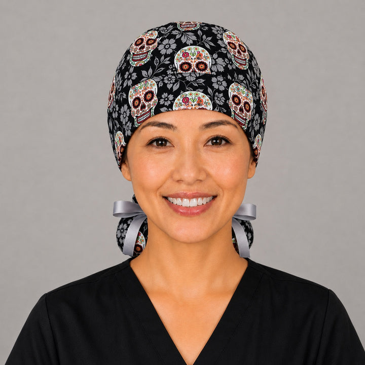 Sugar skull Halloween ponytail scrub cap left side view with full ponytail pouch of Day of the Dead skulls on black floral background and long gray ribbon ties