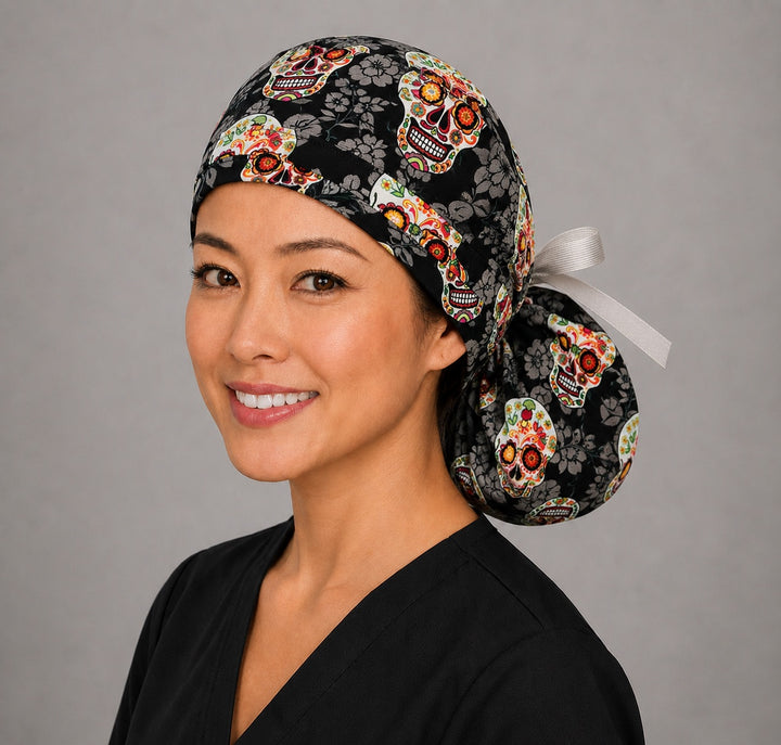 Sugar skull Halloween ponytail surgical cap left three-quarter angle on smiling model showing Day of the Dead skulls on black floral background with gray ribbon bow