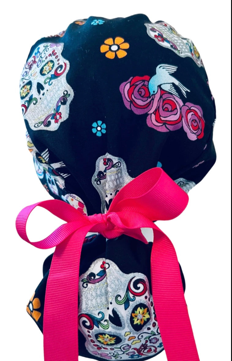 Sugar skull ponytail scrub cap back close-up showing gathered pouch of silver Day of the Dead skulls, roses and a bird on black with hot pink grosgrain ribbon bow