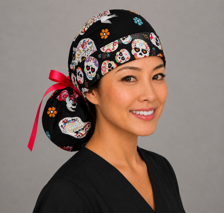 Woman wearing sugar skull ponytail scrub cap, front view smiling with colorful floral sugar skulls and birds on black with sugar-skull band and pink ribbon tie