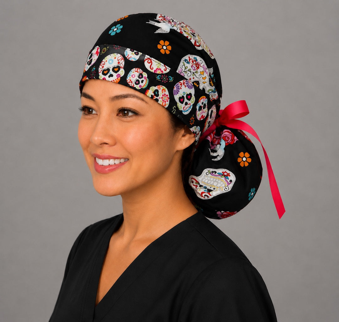 Sugar skull ponytail surgical cap right three-quarter angle on smiling model showing gathered pouch of colorful skulls, roses and birds on black with pink ribbon bow