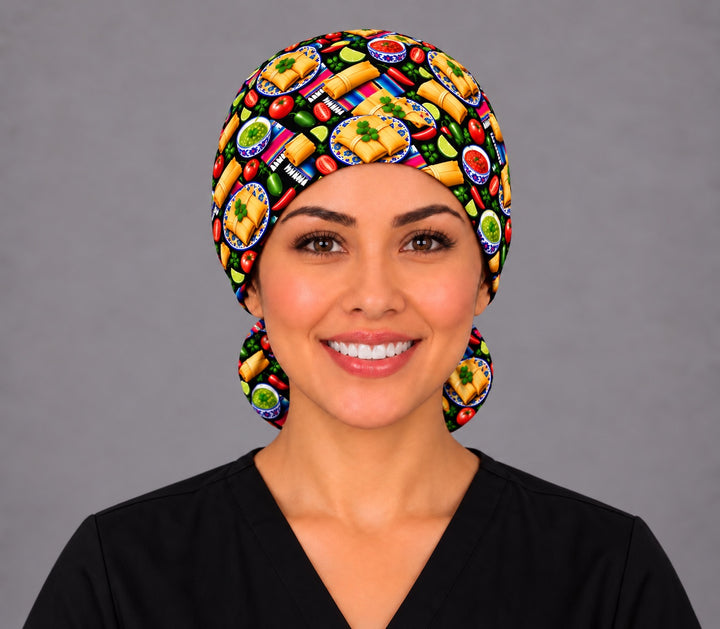 Smiling Latina nurse in black scrubs wearing a Mexican tamale ponytail scrub cap, front view with tamales on Talavera plates across the band and the pouch peeking out below