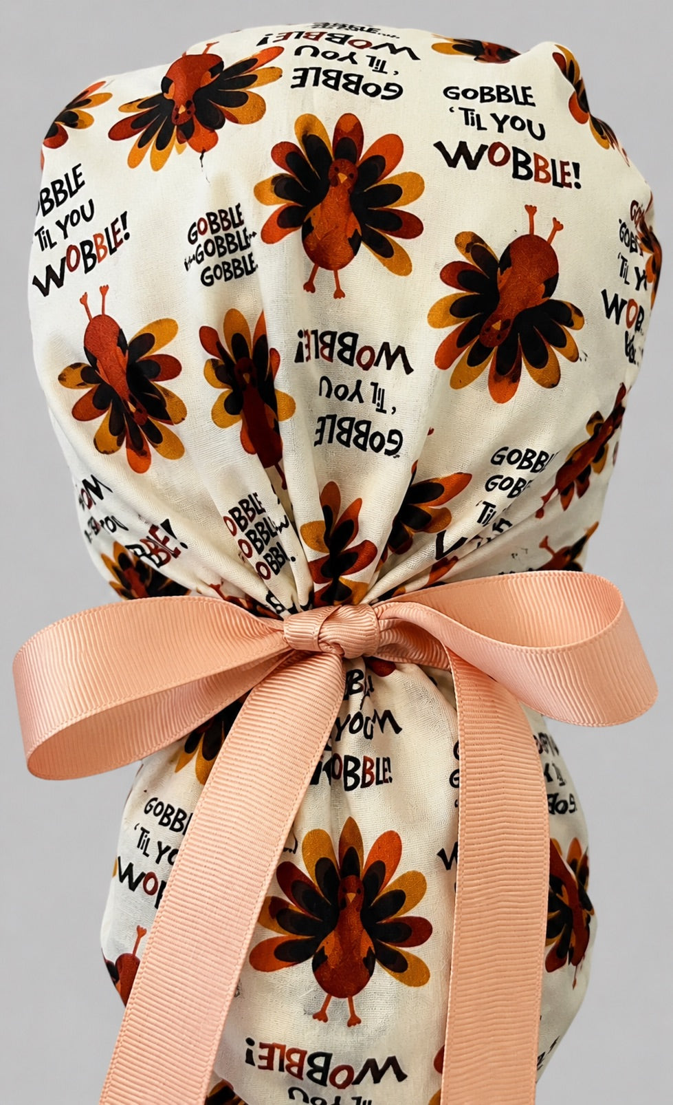 Woman wearing Thanksgiving turkey ponytail scrub cap, front view smiling with turkeys and Gobble Til You Wobble text on cream with pumpkin-spice band and peach ribbon tie