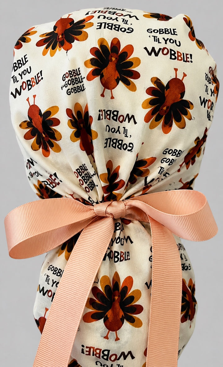Woman wearing Thanksgiving turkey ponytail scrub cap, front view smiling with turkeys and Gobble Til You Wobble text on cream with pumpkin-spice band and peach ribbon tie