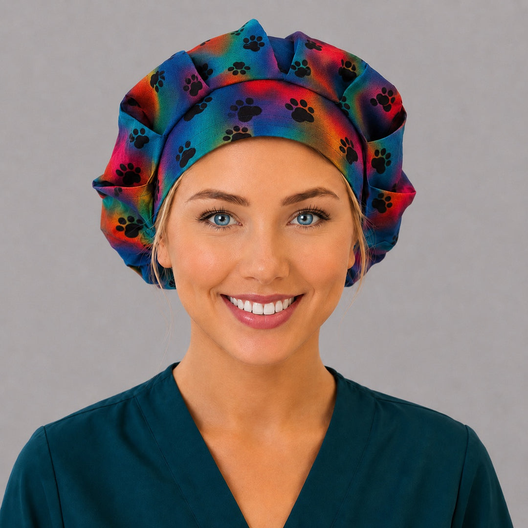 Woman wearing a tie-dye paw print bouffant scrub cap, front view — black paws on rainbow watercolor, adjustable women&
