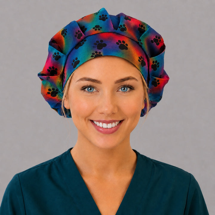 Woman wearing a tie-dye paw print bouffant scrub cap, front view — black paws on rainbow watercolor, adjustable women&