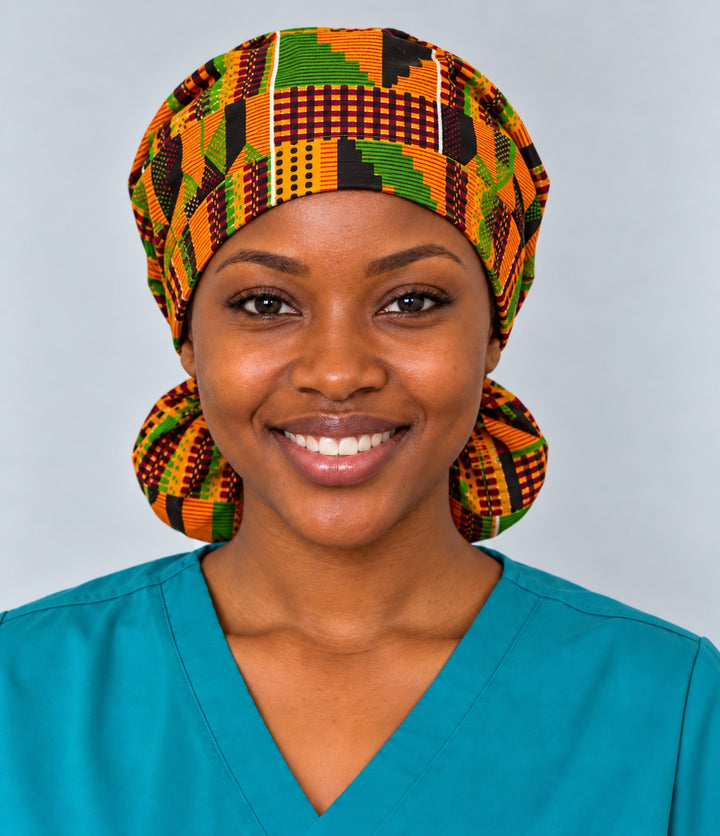 Front view of a smiling nurse in an XL Traditional Kente ponytail scrub cap, with orange, green and maroon Ghana patchwork blocks across the forehead band