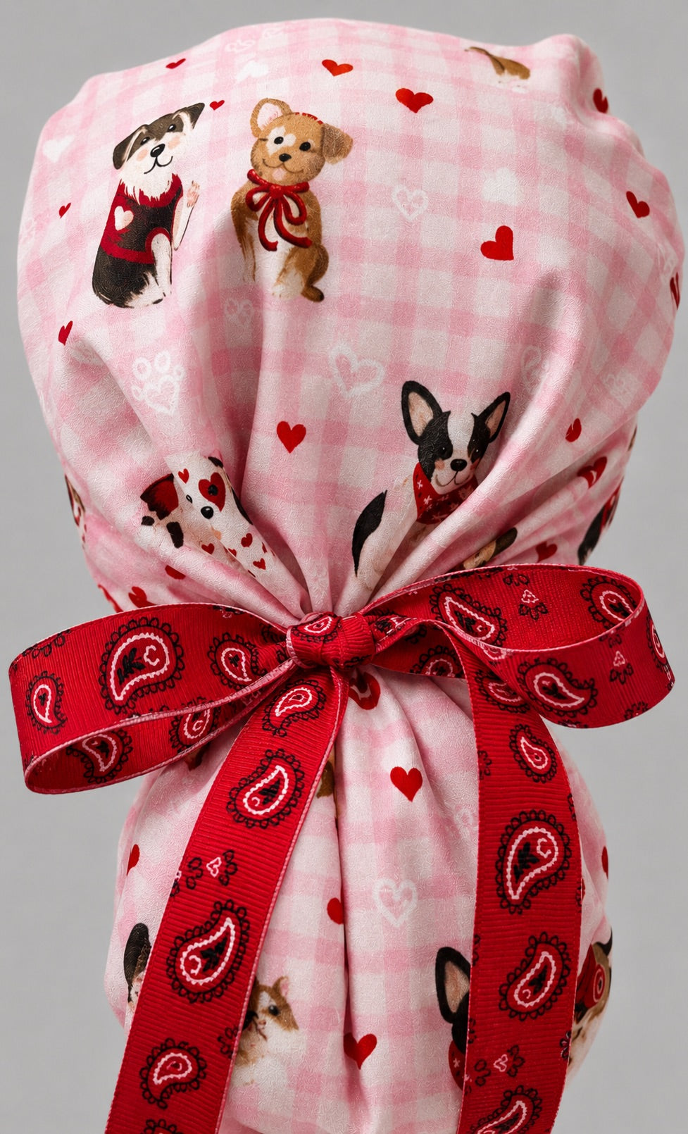 Woman wearing Valentine dogs ponytail scrub cap, front view smiling with puppies, hearts and bows on pink gingham with red bandana band and red ribbon tie
