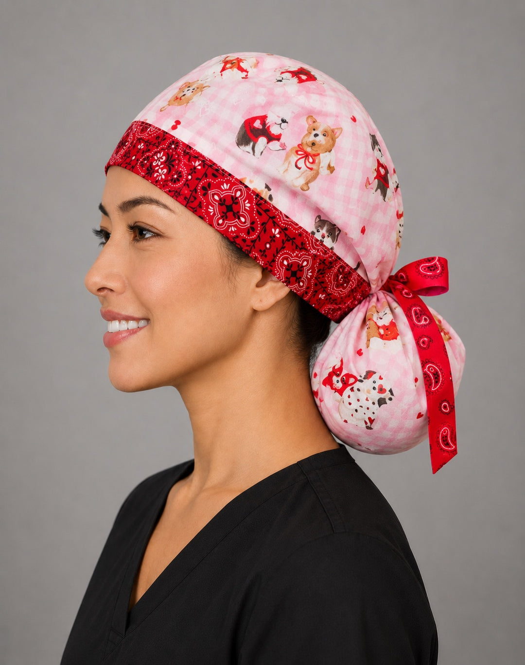 Valentine dogs ponytail scrub cap right three-quarter angle on smiling model showing puppies, hearts and bows on pink gingham with red bandana band and red ribbon bow