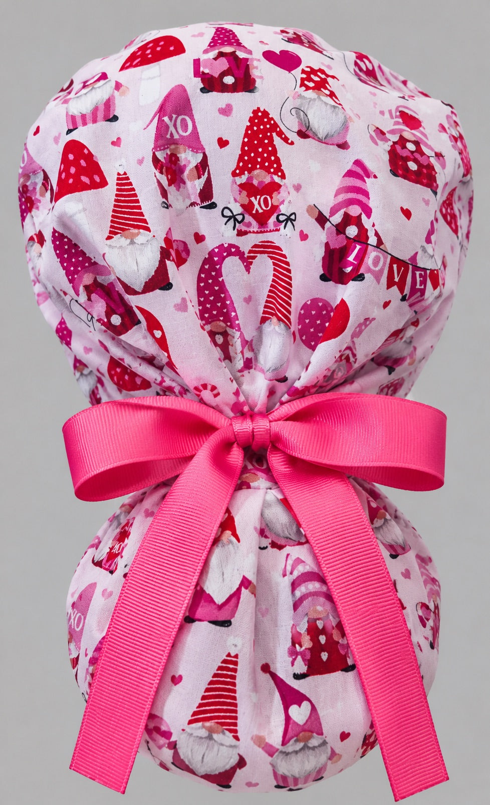 Woman wearing Valentine gnomes ponytail scrub cap, front view smiling with red and pink gnomes and hearts on pink with red heart band and pink ribbon tie