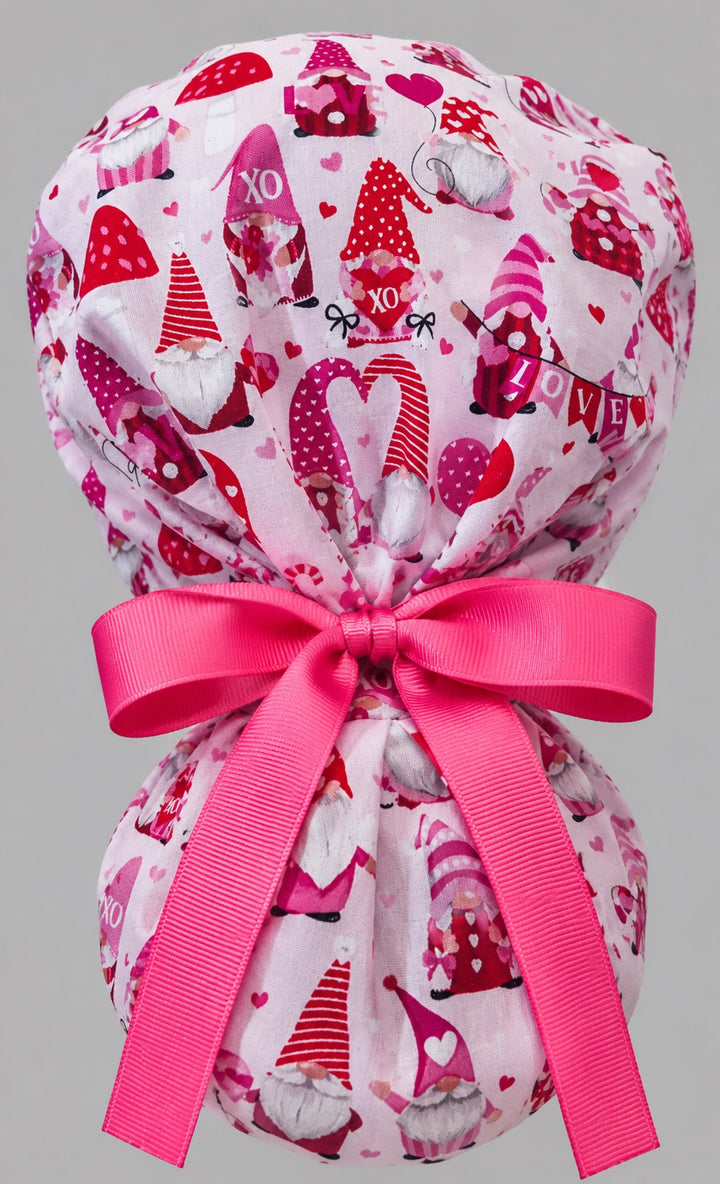 Woman wearing Valentine gnomes ponytail scrub cap, front view smiling with red and pink gnomes and hearts on pink with red heart band and pink ribbon tie