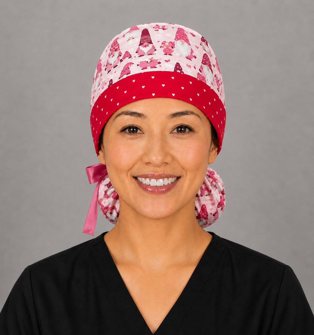 Valentine gnomes ponytail nurse cap left three-quarter angle on smiling model showing red and pink gnomes, hearts and LOVE banner with red heart band and pink ribbon bow
