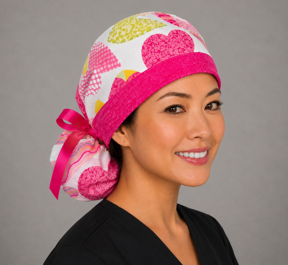 Woman wearing Valentine's hearts ponytail scrub cap, front view smiling with pink, yellow and lime hearts on white and hot pink band with pink ribbon tie