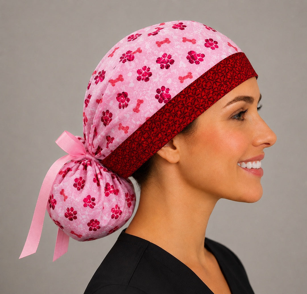 Woman wearing Valentine puppy paws and bones ponytail scrub cap, front view smiling with pink and red paw prints, dog bones and red band with pink ribbon tie